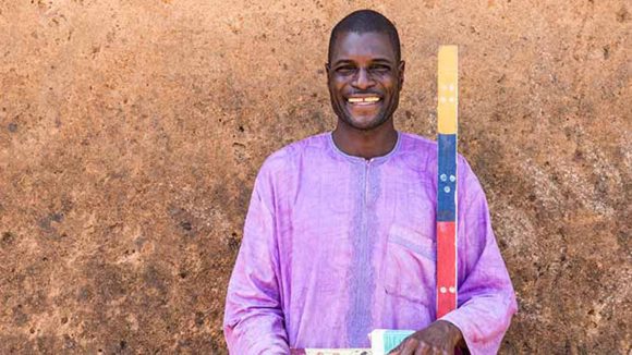 Ayuba Auta, Mectizan distributor in Bugai, stands against a wall with a dosing pole. He is wearing purple and smiling.