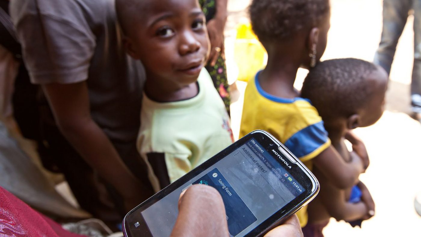 An eye health worker holds a mobile phone to record data, as a boy in the background looks on.