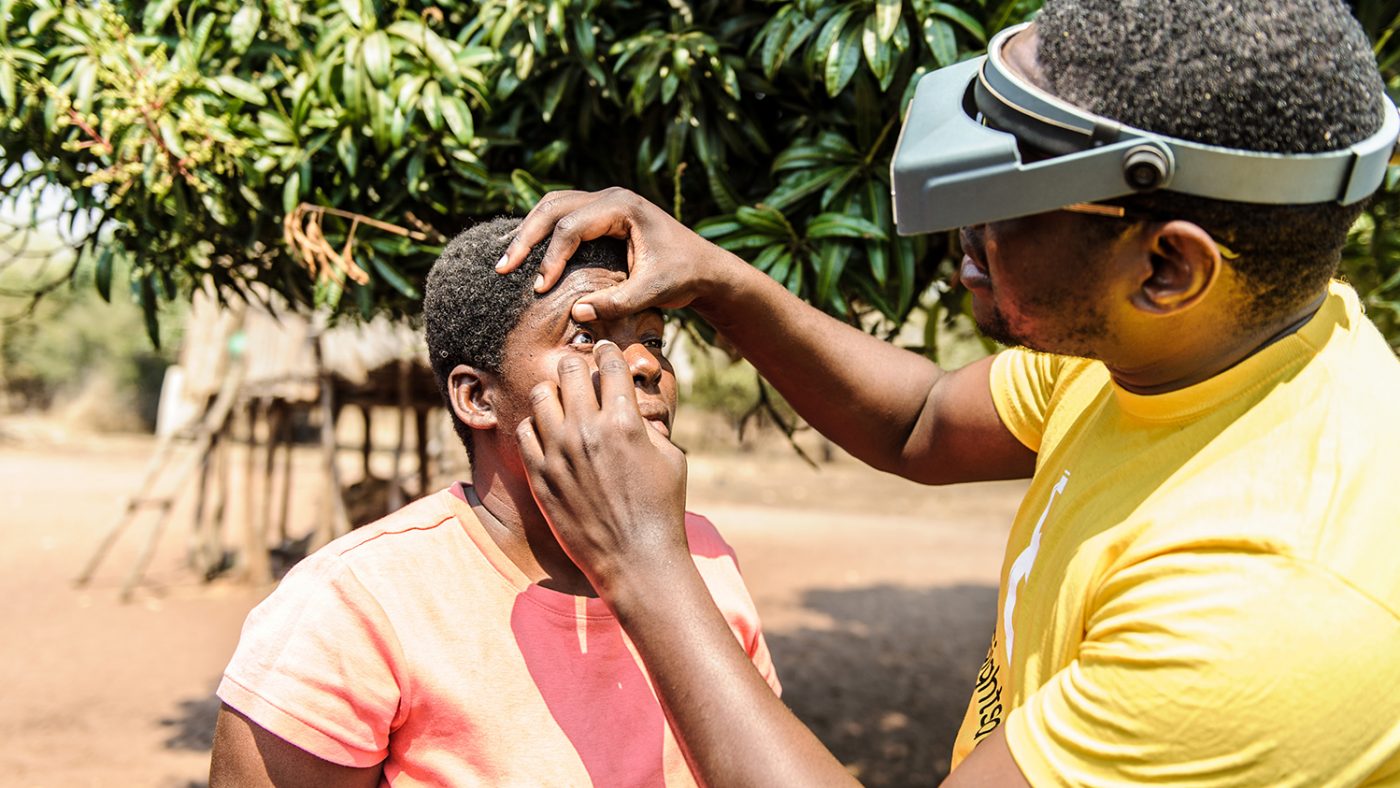 A man in a Sightsavers tshirt examining a lady's eyes for trachoma.
