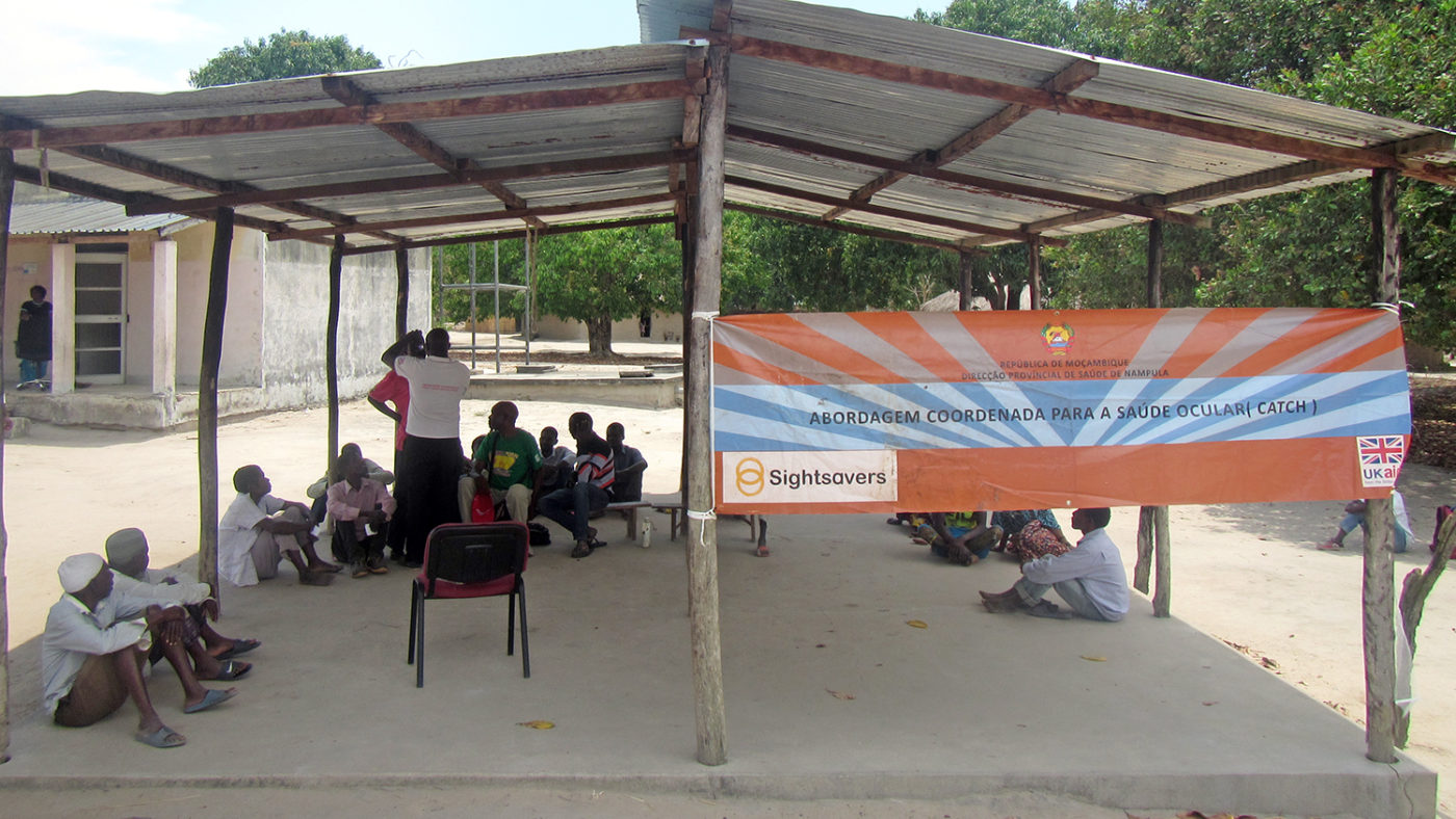 A temporary structure with people sitting underneath, with a sign featuring the Sightsavers logo.