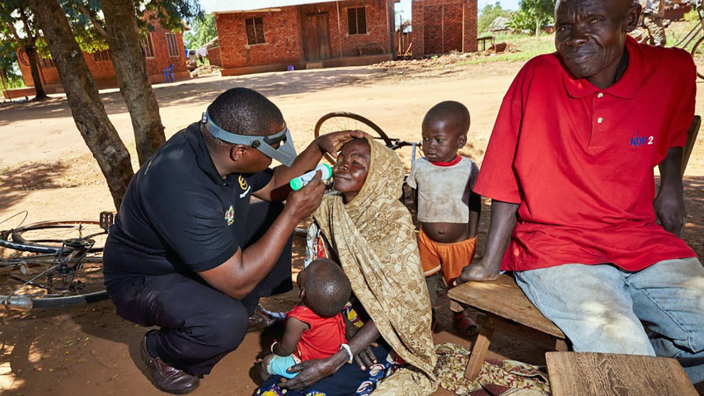 An eye health worker examines a woman's eyes, with her family gathered round her.
