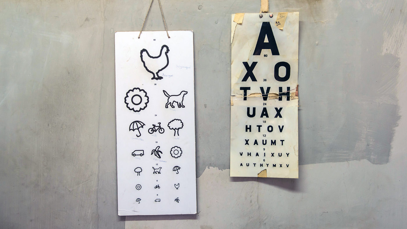 Two eye test charts hang on a wall. One has symbols of animals and objects. The other chart is a Snellen chart with letters.