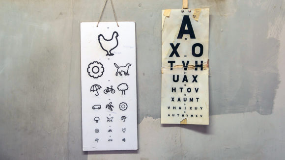 Two eye test charts hang on a wall. One has symbols of animals and objects. The other chart is a Snellen chart with letters.
