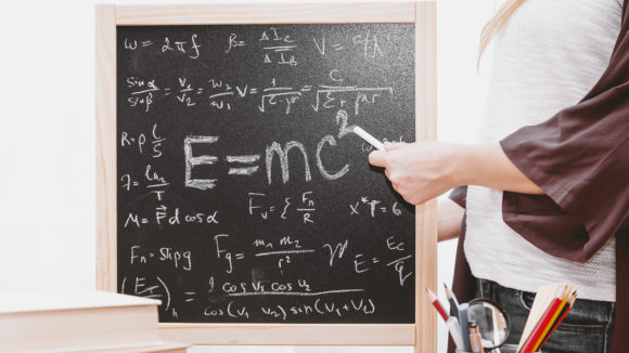 A man using a piece of chalk to point at a blackboard, which shows the text 'E=MC squared'.