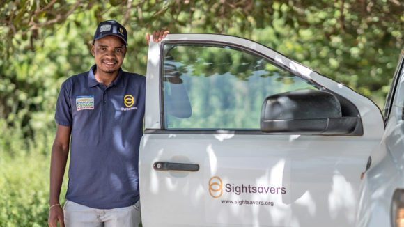An eye health worker wearing a purple t-shirt with the People's Postcode Lottery and Sightsavers logos, standing next to a van with the Sightsavers logo on the door.