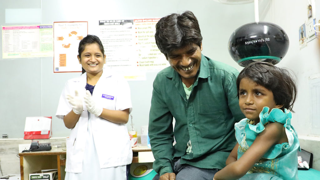Rukmani sits in hospital after her cataract operation. Her father and a nurse smile behind her.