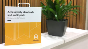 The Sightsavers accessibility pack box: a white and yellow box with handle, featuring the words 'Accessibility standards and audit pack, produced by Sightsavers'.
