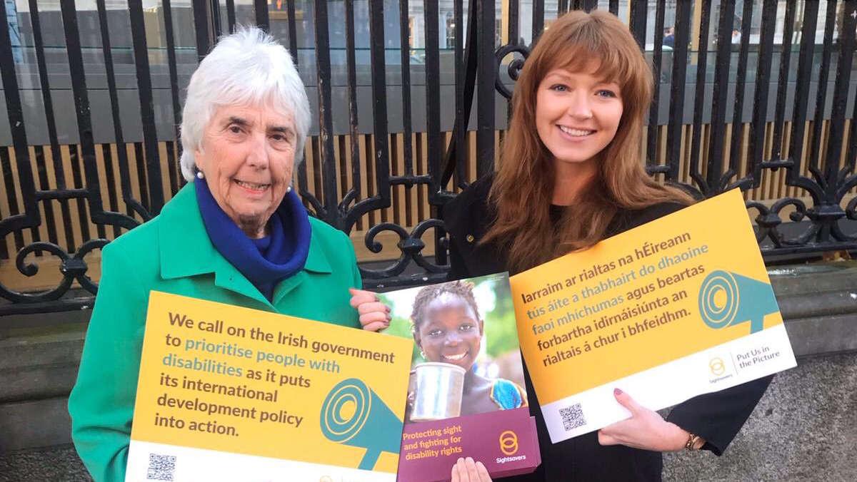 Two women stand together, smiling. They hold petition signs.