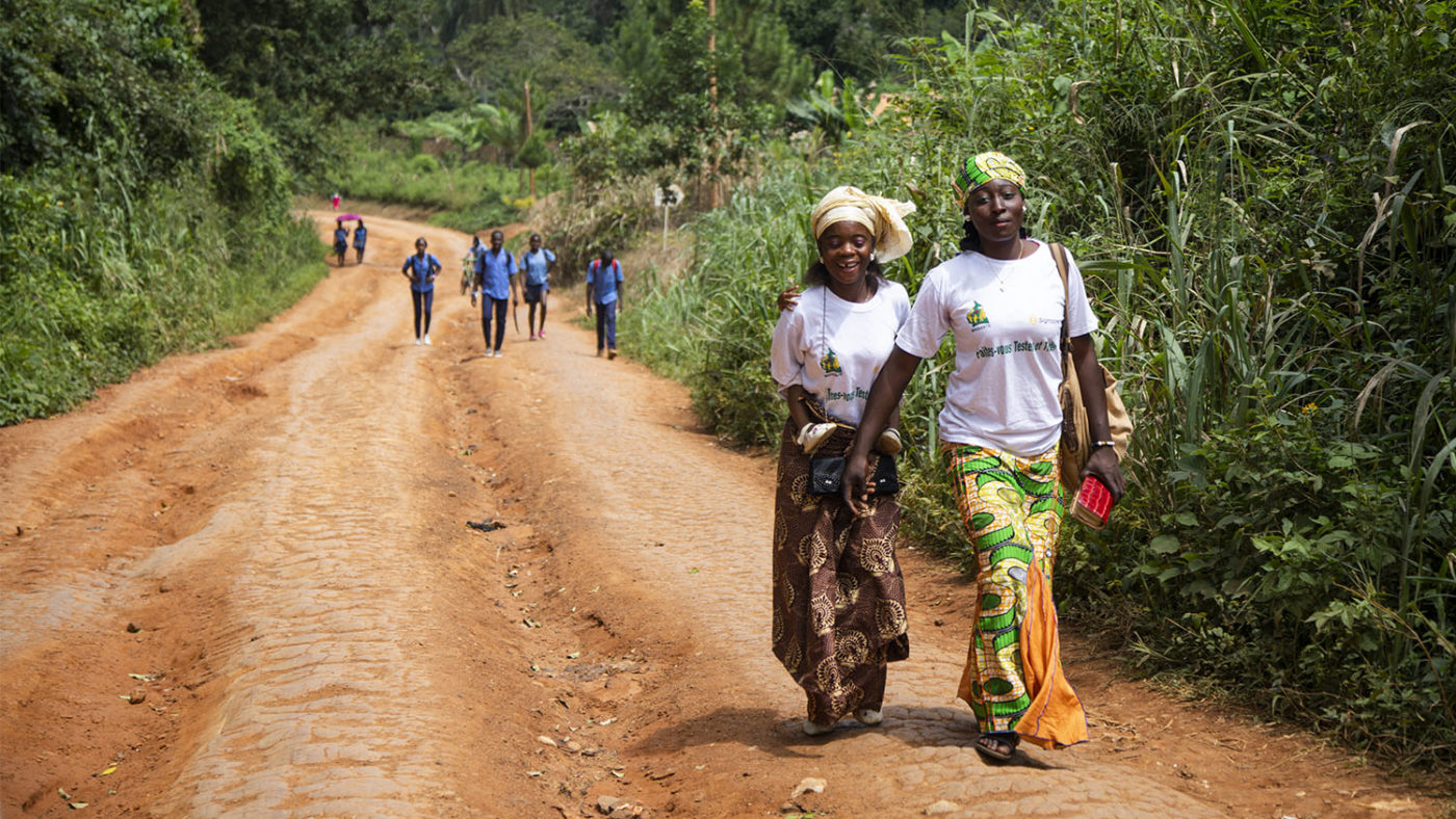 Two women walk along a dirt track surrounded by greenery.