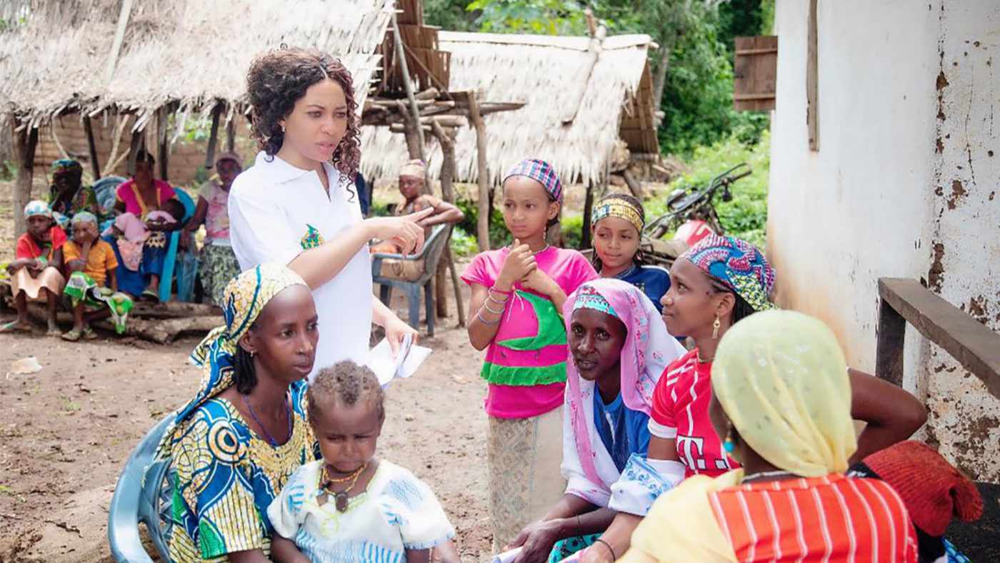 A woman speaks to a group of women and children seated outside,