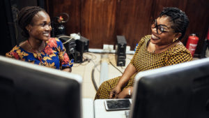 two women sit behind a computer, laughing.