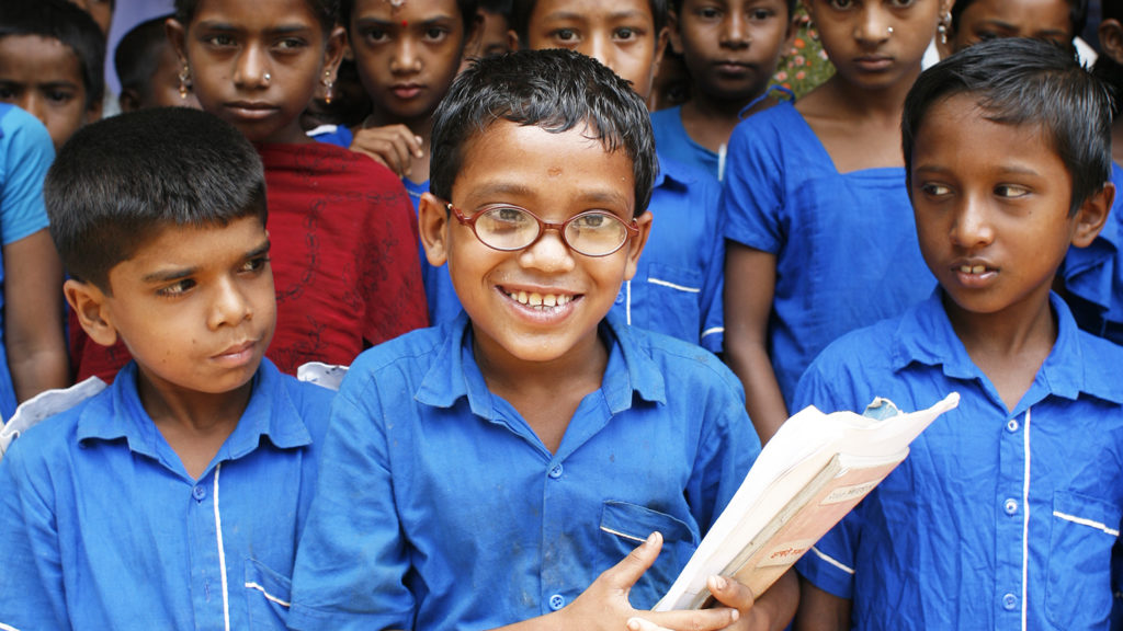 A young boy in school uniform, holding paperwork and smiling.