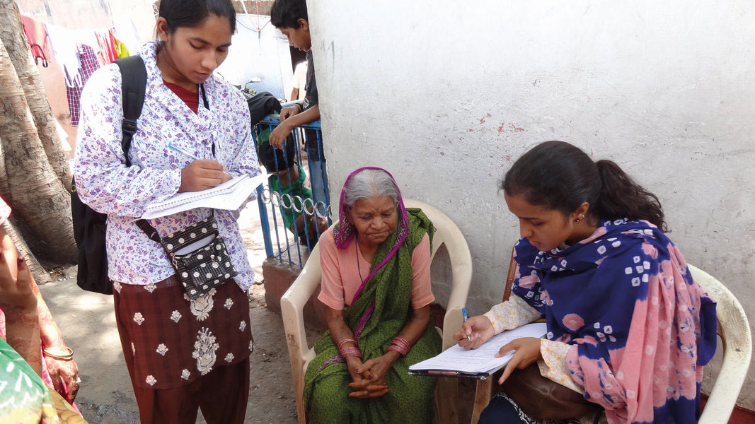 Two young women holding clipboards make notes with a pen, and are shown next to an elderly woman who is seated outside in Bhopal, India.