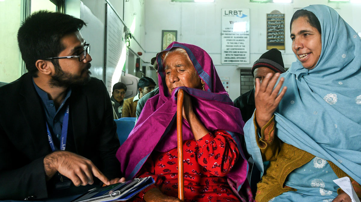 An elderly woman and a younger woman sit next to a man who holds a clipboard, in a hospital waiting area.