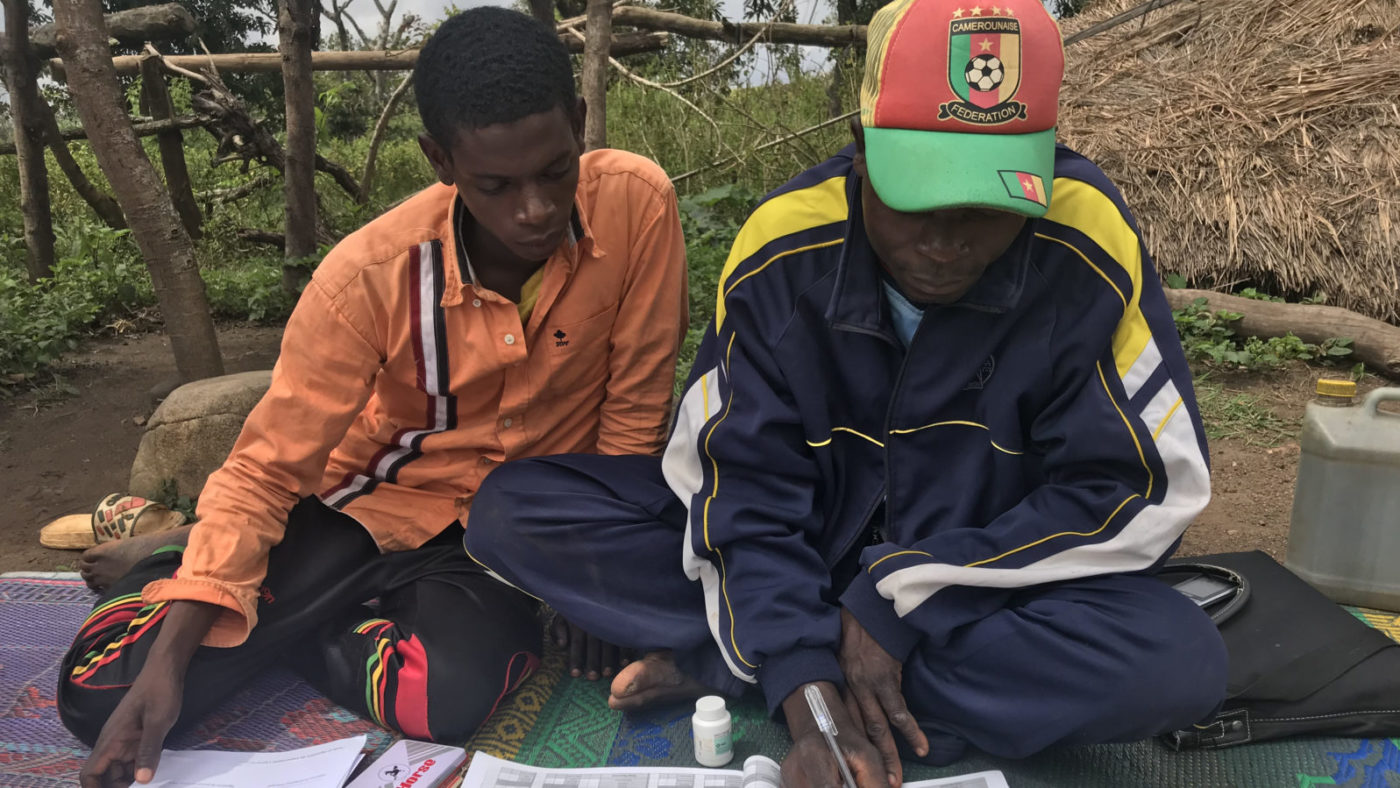 Two men sit side by side outside, one looking at the other filling in a form.