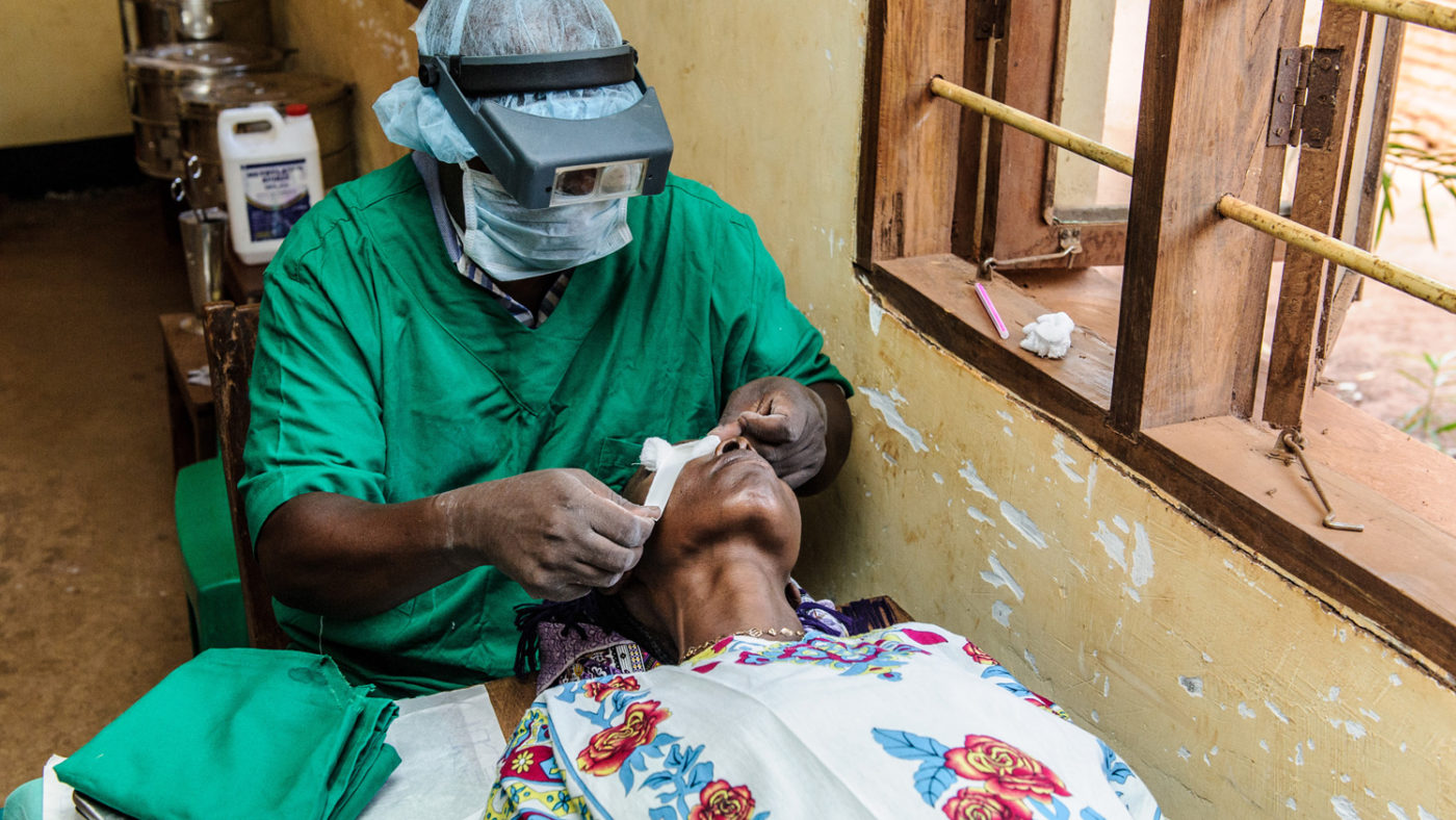 A doctor wearing green scrubs with a visor applying a dressing to a patient's eye.