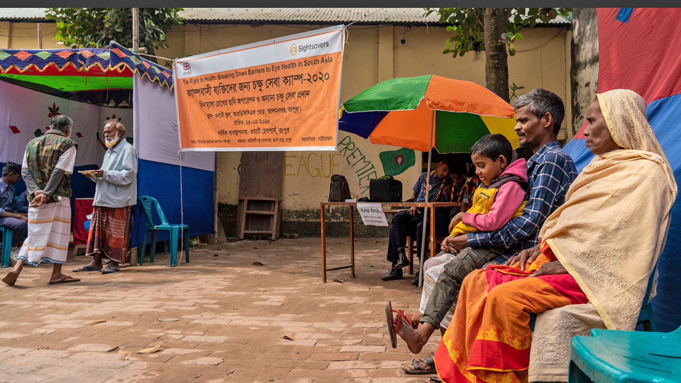A woman, man and child sit next to a banner, promoting Sightsavers free eye health screening for the Bihari community.