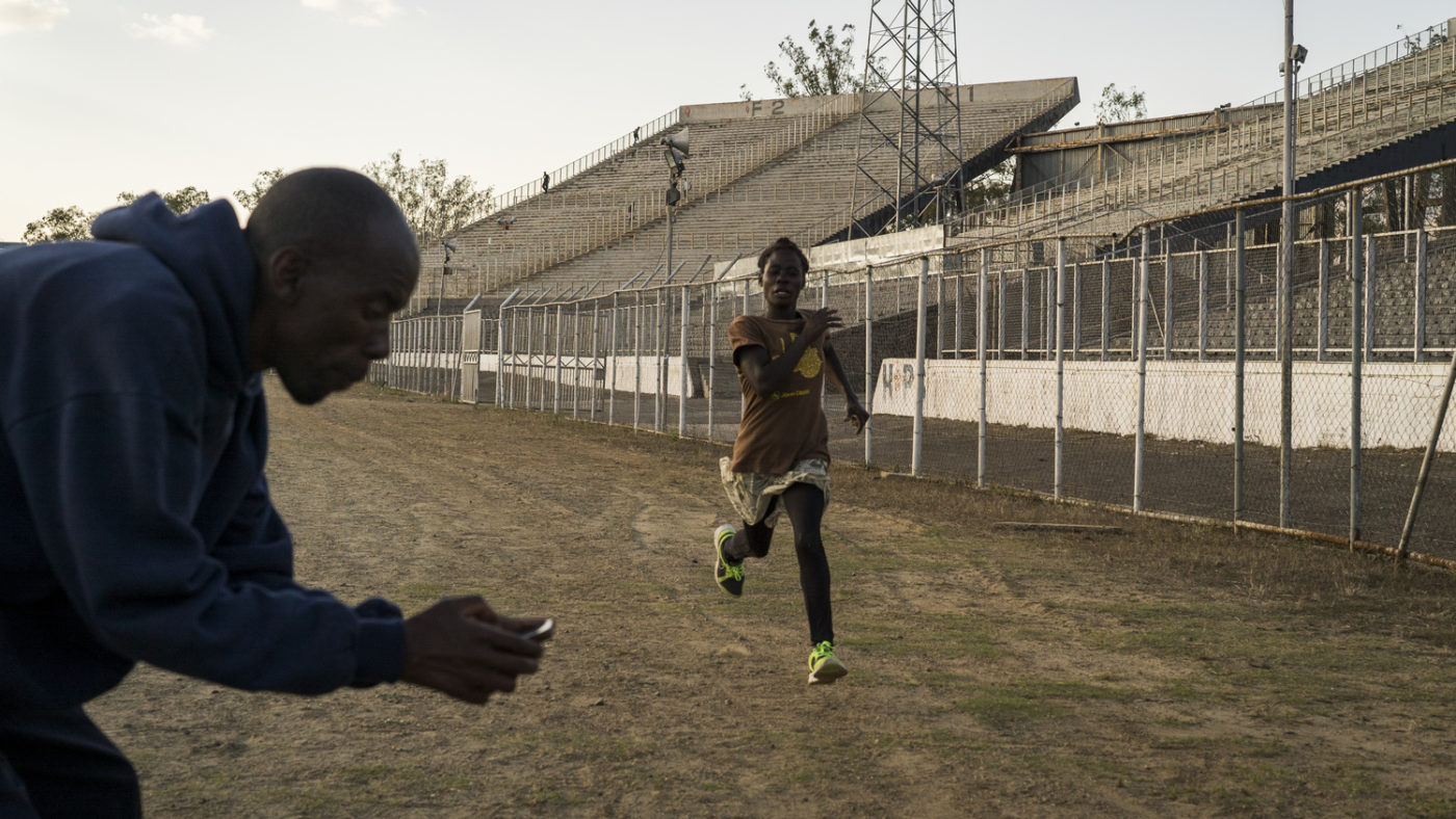A young woman runs on a training track while a man times her with a stopwatch.