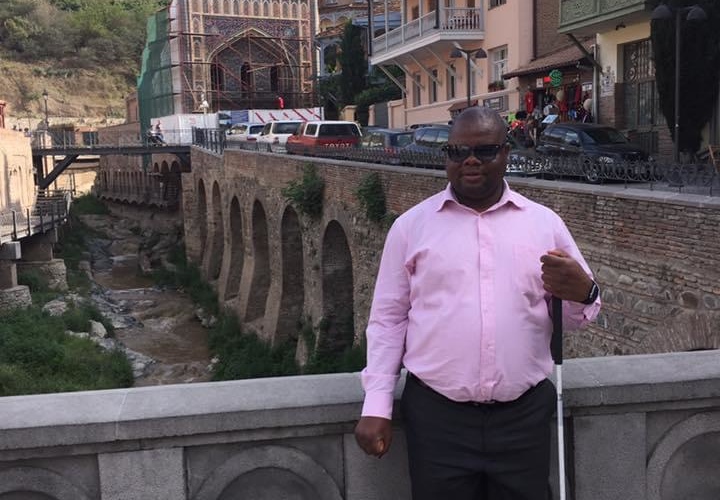 A man standing by a bridge, holding his white cane.