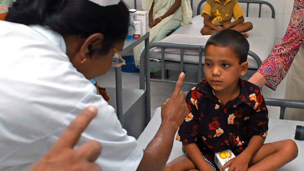 A young boy sits on a hospital bed, looking at a nurse with a finger held up.