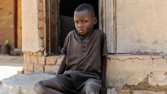 A young boy, Bretty, looks at the camera while sat on the ground outside a doorway.