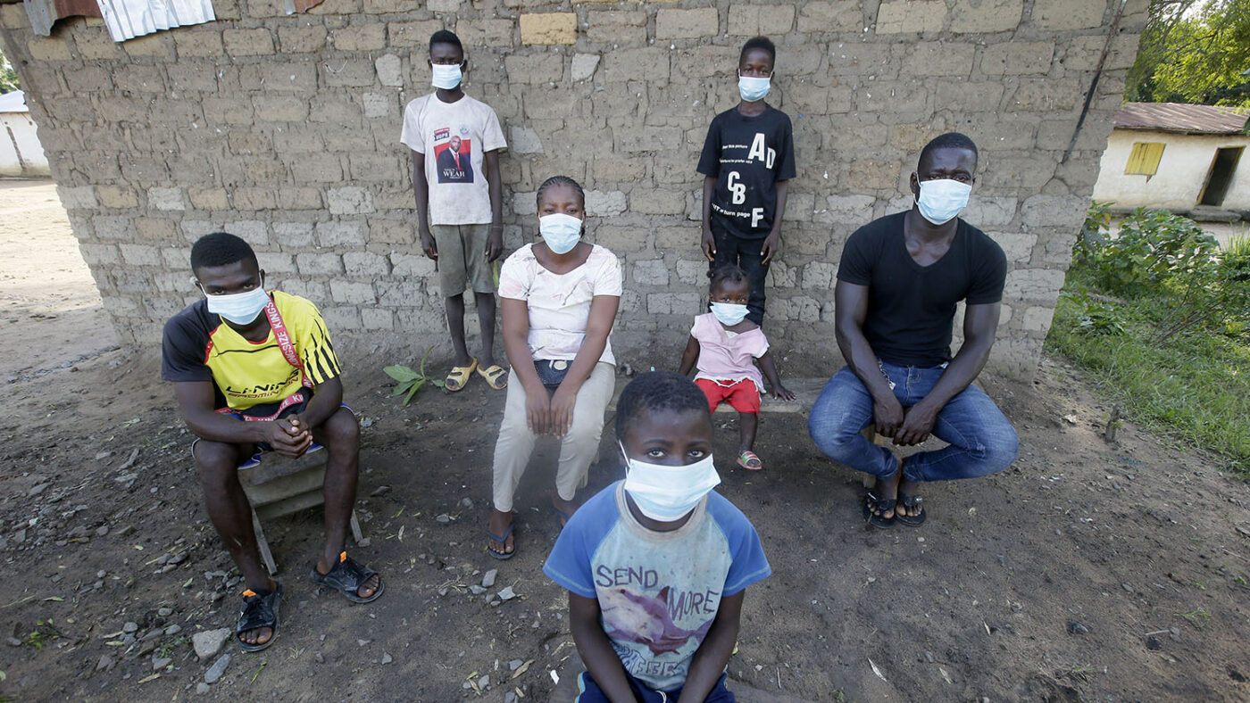 A family, all wearing masks, sit for a social distanced portrait outside their home.