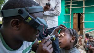 A man wearing magnifying glasses over his eyes shines a torch into a toddler's eye during a trachoma survey.