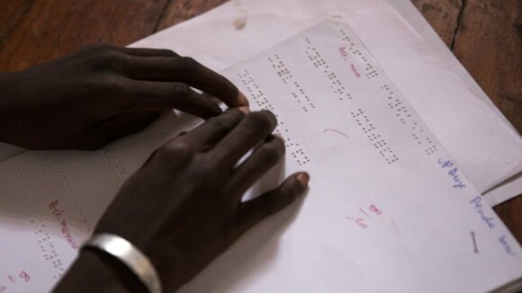 A girl, wearing a silver armband, reads braille text. Only the girls' hands are shown.