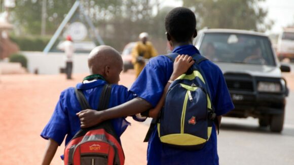 Two schoolboys, arm in arm, walk down a dirt track by the side of a road.