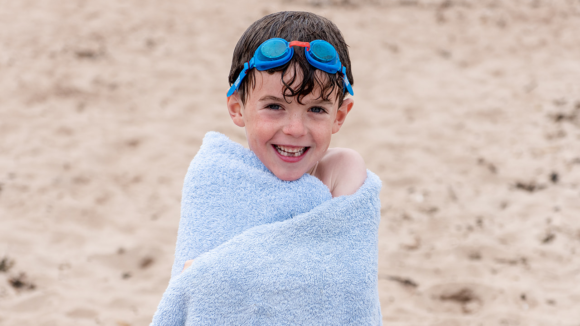 Six-year-old Bertie stands on a beach wrapped in a towel and wearing goggles on his forehead, while smiling broadly.