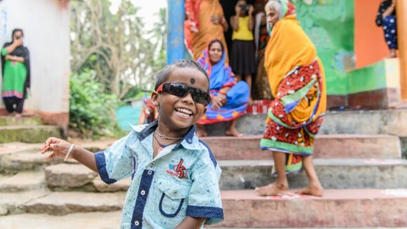 A young boy, Omm, smiles playing outside home wearing protective sunglasses after surgery.