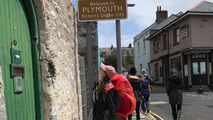 A young woman, Lily, stares up at a sign saying 'Welcome to PLymouth' while wearing a cap and large rucksack.
