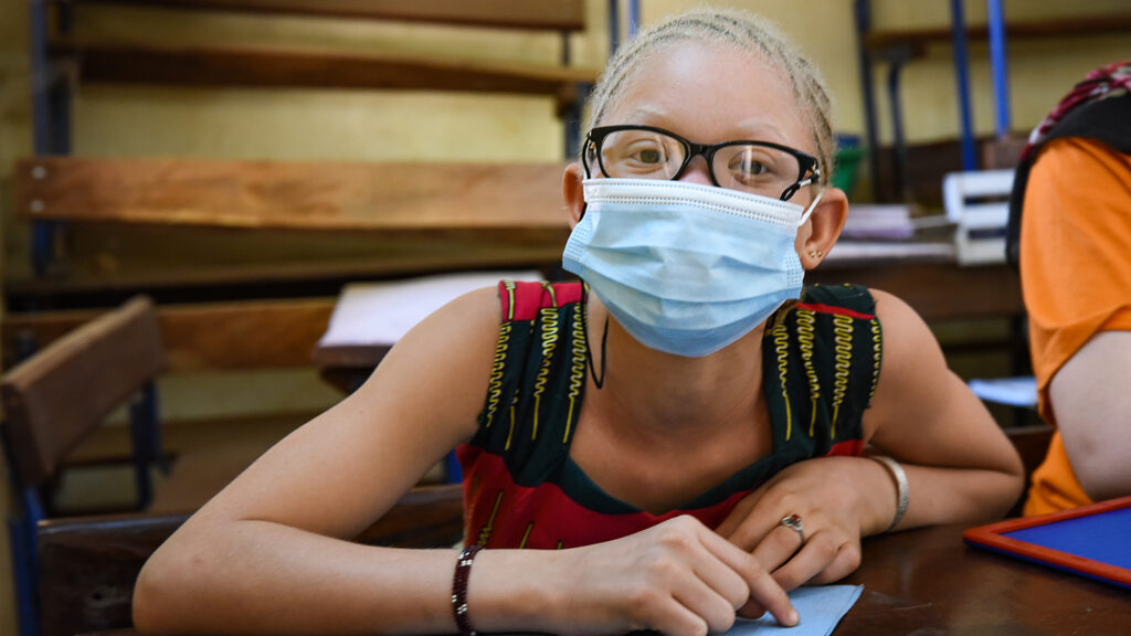 A girl, who has albinism and is wearing glasses and a mask, sits in a classroom.
