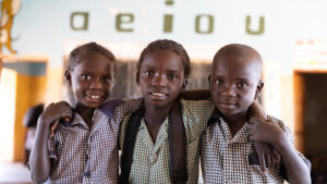 Three young children huddle together at an inclusive school in Zambia. Behind them on the wall are the vowels A, E, I, O, U.
