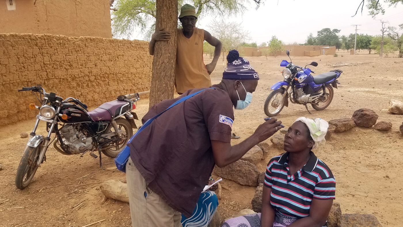 An eye health worker checks the eyes of a woman sitting on a low wall in a dusty setting.