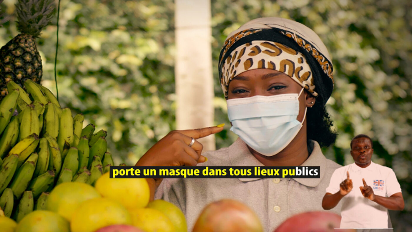 An image showing a woman at a market, with piles of different fruits in the foreground. The woman is pointing to a mask that she is wearing over her nose and mouth.