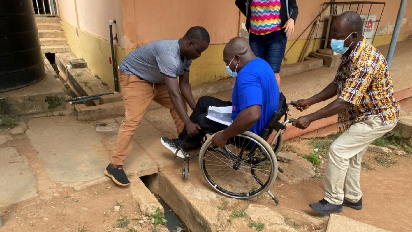 Three men, including one wheelchair user, navigate uneven terrain in the hospital grounds