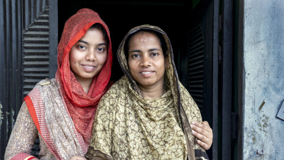 in Bangladesh, two women stand with their arms round each other outside their home.