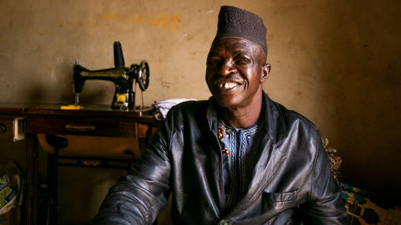 A man sitting in front of a sewing machine smiles at the camera.