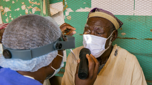 Sina wears a mask while an eye health worker shines a torch into his eyes to check for trachoma.