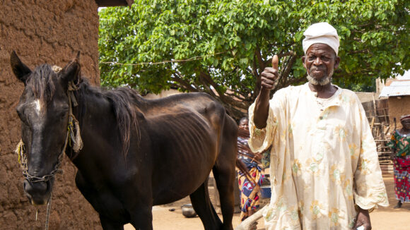 Sonbobia stands outside his home with his horse. He holds up his thumb and smiles at the camera.