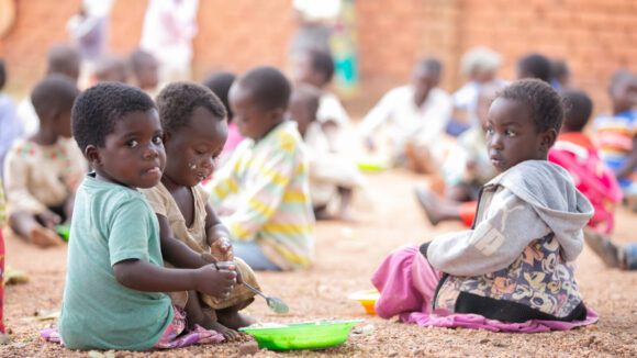 Young children sit together outside eating porridge