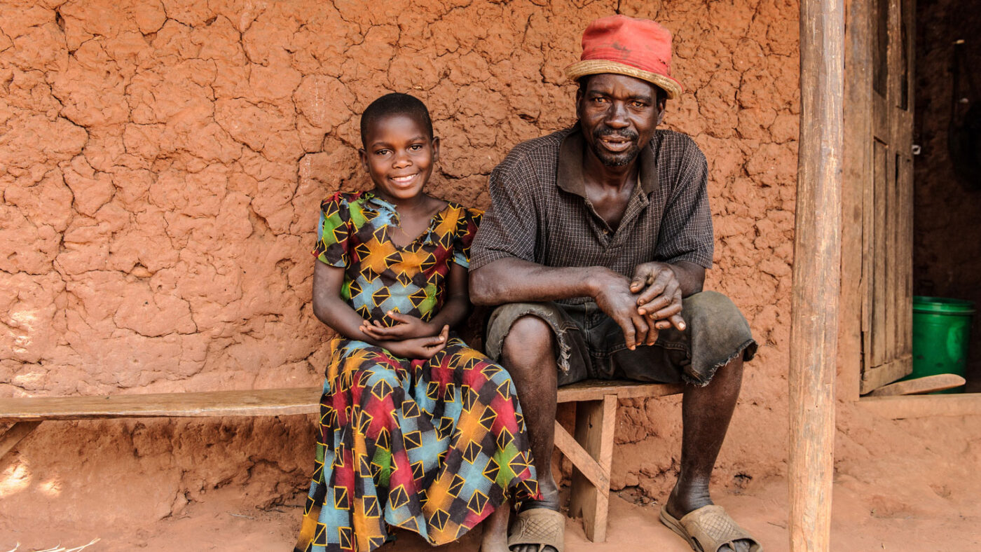 A young girl sits outside smiling on a bench wearing a colourful dress next to her father.