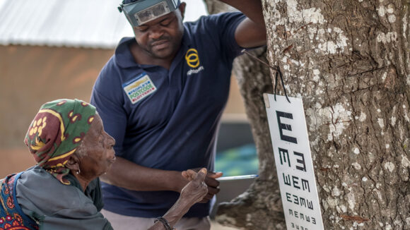 An eye health worker wearing a navy t-shirt points to an eye test chart hanging on a tree, while an older woman looks at the chart.