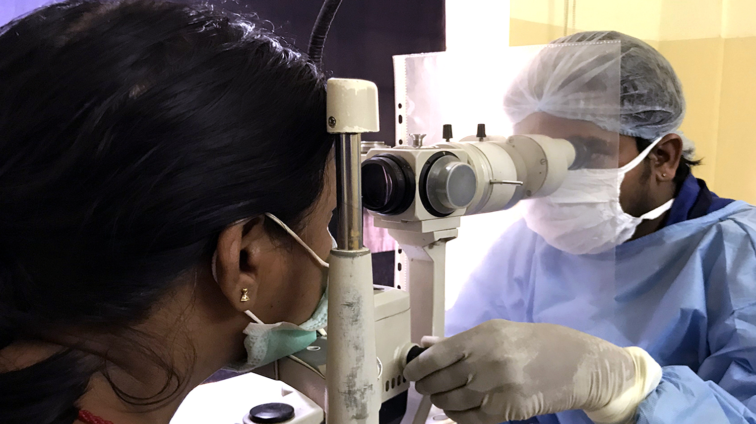 An eye health worker wearing a mask and surgical gown checks a woman's eyes using optical equipment.