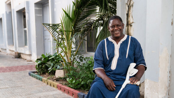 A man smiles at the camera. He's sitting in a chair and holding a white crutch.