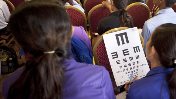 A young woman sitting in a group of people holds an eye test chart.