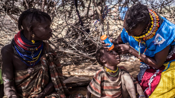 A volunteer wearing a colourful neckpiece and a blue tabbard shines a torch into a boy's eyes to check for signs of eye disease. They're sitting outside under a tree as the boy's mother looks on.