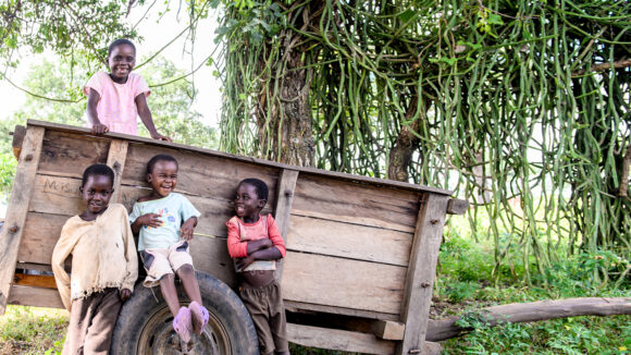 Four children smile and laugh as they sit on a wooden cart amid lush green trees.