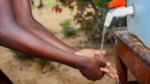 A close-up of a school student washing her hands using water running from a tap in a large water butt.